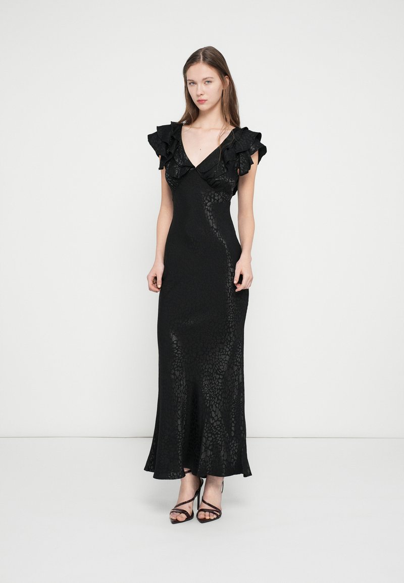 Juoda suknelė su raukiniais ant rankovių, tekstūruotos medžiagos su subtiliu raštu, aptempta silueto ir išplėstais hemu, kartu su juodais bateliais su dirželiais.