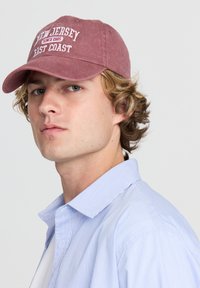 Cappellino da baseball in cotone bordeaux con testo ricamato bianco che recita "New Jersey East Coast dal 1985." Indossato con una camicia a righe blu.