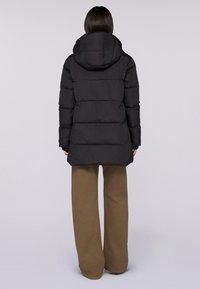 Schwarze Daunenjacke mit Kapuze, ausgestattet mit einem gesteppten Design, verstecktem Reißverschluss und gerippten Bündchen. Über beigefarbenen weit geschnittenen Hosen getragen.