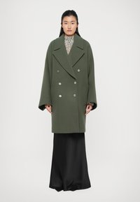 Oversized green wool coat with double-breasted closure, large lapels, and silver buttons. Worn over a long black satin skirt.