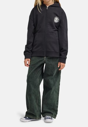 Kind trägt schwarzen Reißverschluss-Hoodie mit Logo, dunkelgrüne weit geschnittene Jeans und marineblaue Turnschuhe vor einfarbigem Hintergrund.