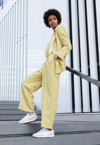 Ljusgul skräddarsydd kostym med vida byxor och matchande kavaj, bärs med vita sneakers, stående på grå trappor mot en modern byggnadsbakgrund.