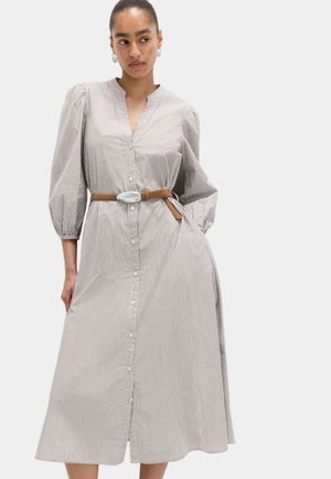 Kobieta ubrana w midi sukienkę na guziki z długim rękawem i paskiem w kolorze brązowym, stojąca z jedną dłonią w kieszeni, patrząca na bok.