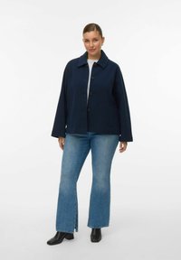 Ženska stoji oblečena v mornarsko modro jakno z gumbi, svetlo modre kavbojke z sprednjimi razporkami, črne gležnjarje in belo majico na belem ozadju.