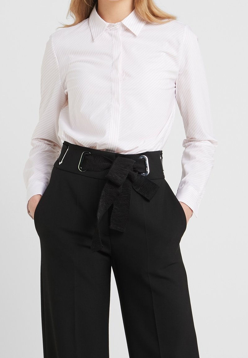 Femme portant une chemise blanche à manches longues boutonnée, rentrée dans un pantalon noir taille haute avec une large ceinture en tissu noire nouée en nœud.