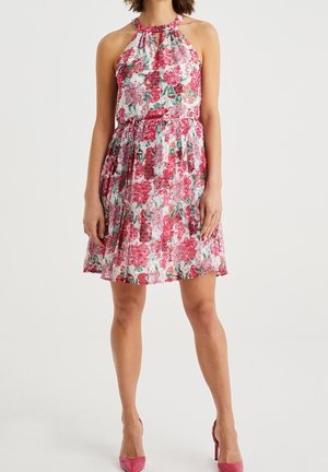 Woman wearing a sleeveless floral dress with a halter neck and pink high heels, standing against a plain white background.