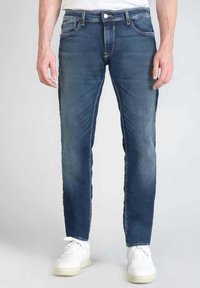 Vaqueros de mezclilla azul oscuro con corte ajustado, que presentan bolsillos tradicionales, costuras en contraste y una ligera textura desgastada. Combinados con zapatillas blancas.