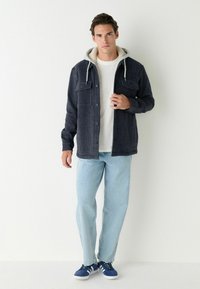 Blau karierter Flanellhemd mit zwei Brusttaschen, grauer Kapuze und weißem Rundhals-T-Shirt darunter. Hellblaue Jeans und blaue Turnschuhe.