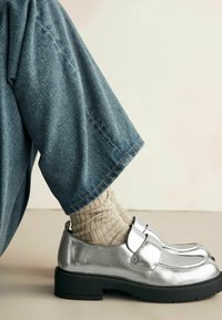 Zapatos de cuero plateado con punta redondeada, suela de goma negra y detalle de hebilla. Combinados con calcetas beige acanaladas y pantalones de mezclilla azules.