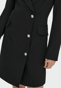 Abito blazer nero su misura con revers, sei bottoni argentati con motivo floreale e due tasche laterali. Tessuto liscio con un design aderente.