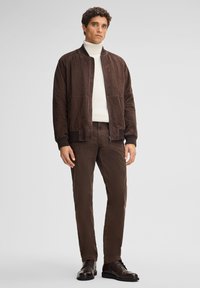 Giacca bomber in suede marrone con polsini e colletto a coste, abbinata a pantaloni marroni e stivali in pelle scura. Texture liscia e vestibilità casual.