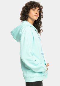 Turquoise hoodie gemaakt van zachte katoen. Voorzien van een zak aan de voorkant, een trekkoord kap en een grafische print. Lange mouwen en een ontspannen fit.