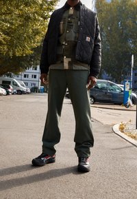 Zwarte gewatteerde jas, camouflage shirt, groene broek en zwart/rode schoenen. Buiten op asfalt met bomen en geparkeerde voertuigen op de achtergrond.