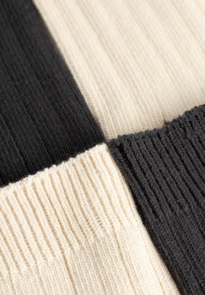 Tuva smalkā režģa adījuma auduma ar mainīgām melnām un krēma krāsas kvadrātu sadaļām, redzama adījuma tekstūra un šuvju detaļas.