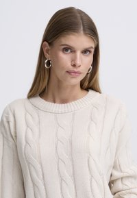 Kremasti džemper s kabelskim uzorkom, okruglim izrezom i rebrastim obrubom. Tkanina je mekana, s teksturiranim uzorkom i opuštenim krojem.