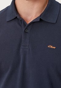 Granatowa koszulka polo z fakturowanym splotem, dwa guziki przy kołnierzyku oraz małe logo s.Oliver w kolorze pomarańczowym na lewej piersi.