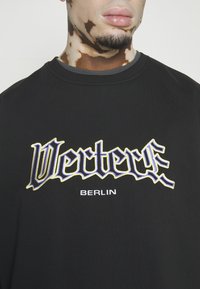 Young man with vitiligo on neck and dark facial hair wearing a black sweatshirt with "Vecterus Berlin" text on the chest.