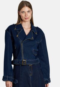 Chaqueta de moto de mezclilla oscura con cintura con cinturón, cierre delantero con cremallera y bolsillos rectangulares. Presenta accesorios en tono dorado y solapas anchas.