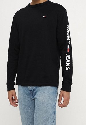 Czarna koszula z długim rękawem z małym logo Tommy Jeans na klatce piersiowej oraz dużym napisem "TOMMY JEANS" i flagą na prawym rękawie, noszona z jasnoniebieskimi dżinsami.