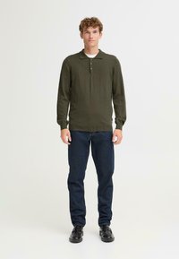 Polo a maniche lunghe verde oliva scuro con colletto a tre bottoni, abbinato a jeans blu scuro e scarpe nere. Design semplice e aderente con polsini a costine.
