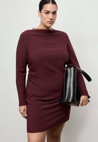 Vestido burdeos de manga larga con escote barco y fruncidos laterales, combinado con un bolso de cuero negro con una correa corta.