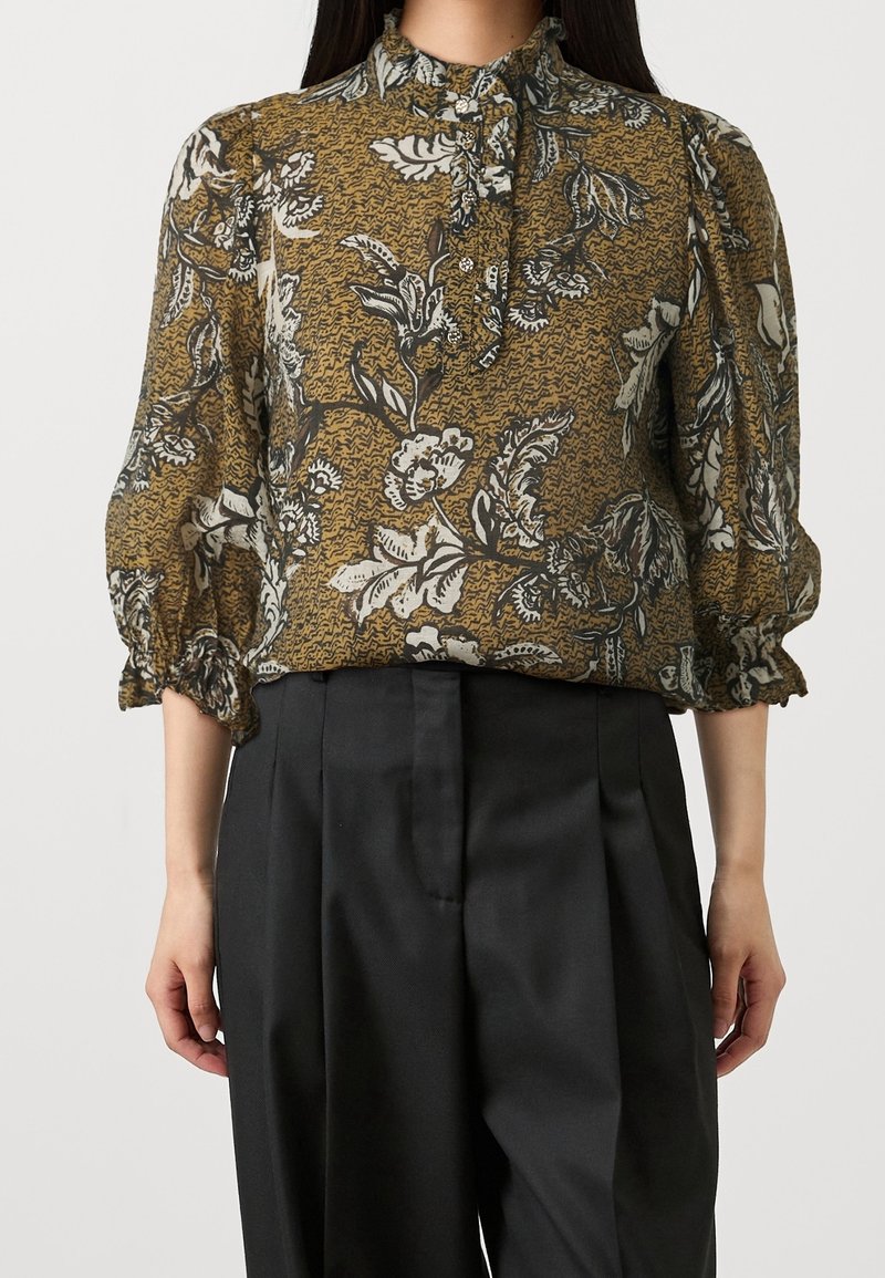Bloemige blouse in mosterd met zwart-witte patronen. Heeft een hoge kraag, knoopsluiting en elastische manchetten. Zacht, doorzichtig stof.