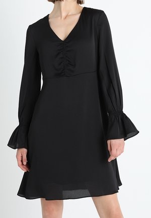 Frau trägt ein knielanges schwarzes Kleid mit langen Ärmeln, die an ausgestellten Manschetten gerafft sind, und einem gerafften V-Ausschnitt.