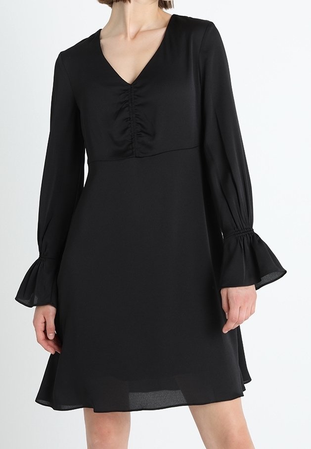 Femme portant une robe noire mi-longue avec des manches longues froncées aux poignets évasés et un décolleté en V froncé.