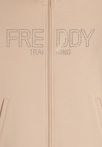 Felpa con zip beige con scritte in strass "FREDDY TRAINING". Presenta due tasche laterali e una consistenza del tessuto liscia.