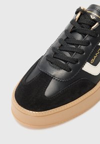 Svart lær- og semsket skinnsneaker med en tan gummisåle, teksturerte detaljer og svarte snører. Logo i kremfarge på siden.