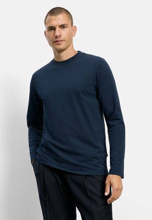Mann mit kurzen Haaren, der ein dunkelblaues langärmeliges Hemd und schwarze Hose trägt, steht mit einer Hand in der Tasche vor einem einfarbigen Hintergrund.