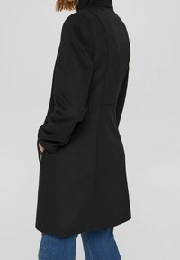 Manteau en mélange de laine noire avec une silhouette ajustée, un col haut et des poches latérales zippées. La surface texturée et la finition douce ajoutent de la profondeur.