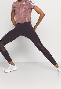 Maglietta cropped Adidas color borgogna con un motivo testurizzato e strisce bianche. Leggings attillati viola scuro con pannelli in rete, abbinati a scarpe da ginnastica bianche.