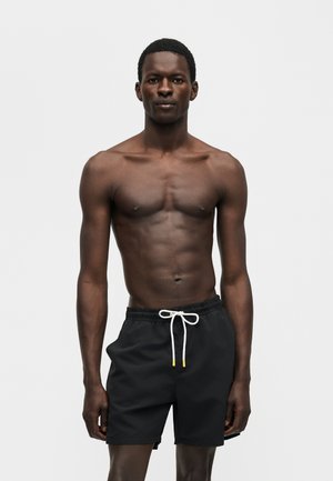 Athletic Black man standing, wearing black swim shorts with white drawstring, against plain white background.