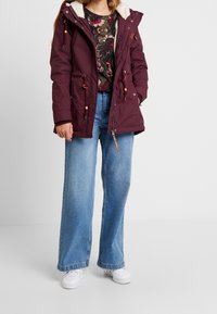 Frau in einer weinroten Kapuzenjacke mit Zugband, floral gemustertem Hemd, hellblauen weit geschnittenen Jeans und weißen Turnschuhen, die vor einem einfarbigen Hintergrund steht.