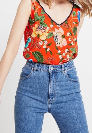 Mujer con vaqueros azules de tiro alto y una camiseta sin mangas roja con grandes estampados florales coloridos y ribete negro en el escote.