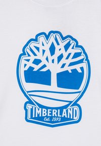Valge t-särk, millel on sinine ümmargune logo stiliseeritud puuga ja tekst "TIMBERLAND Est. 1973" rasvases kirjas.