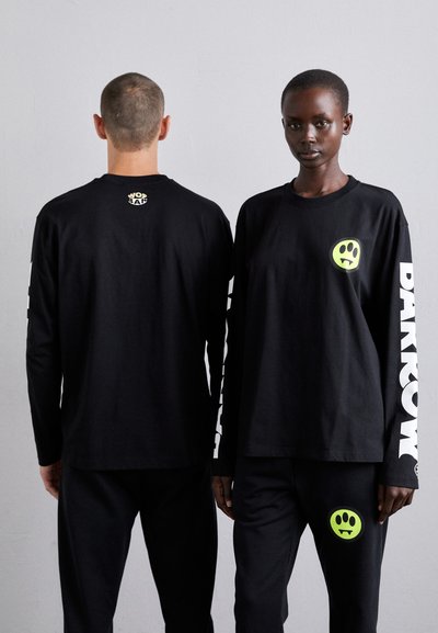 Deux modèles portent des t-shirts noirs à manches longues assortis avec du texte blanc et un graphique de smiley vert. Ils les associent avec des pantalons noirs.