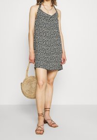 Robe noire avec motif floral blanc, design à col halter, tissu léger, assortie à un sac rond en paille tissée et des sandales à lanières brunes.