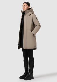 Woman standing, wearing a beige hooded winter coat, black leggings, and tall black boots against a plain light background.