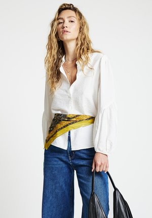 Femme aux longs cheveux bouclés portant un chemisier blanc, un jean bleu, une ceinture écharpe à motifs jaunes, des colliers superposés, et tenant un sac à main plissé noir.