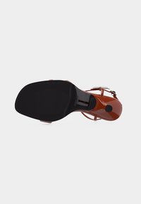 Κόκκινο πέδιλο με ψηλό τακούνι, μαύρη σόλα και λουράκι στον αστράγαλο, εμφανιζόμενο από την κάτω πλευρά σε λευκό φόντο.