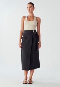 Črna zavita krila z pasom, ki ima stransko žep. Model nosi bež rebrast top in črne ravne sandale. Gladka tekstura tkanine.