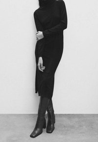 Robe noire à col roulé avec manches longues, dotée d'une fente latérale. Associée à des bottes montantes à talons carrés et finition mate.