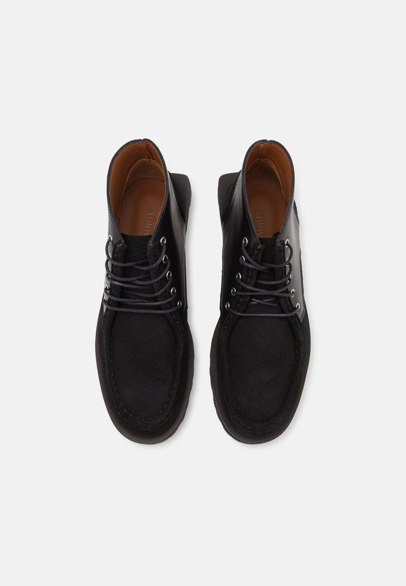 Bottines en cuir noir avec texture en daim, bout rond et œillets en métal pour les lacets. Détails intérieurs marron. Accents cousus.