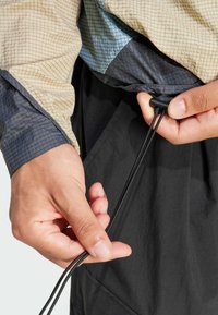 Chaqueta ligera con un patrón de cuadrícula en beige y azul, con un acento en gris oscuro. La mano izquierda ajusta un cordón en el dobladillo.