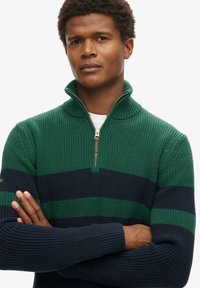 Zielono-granatowy sweter z dzianiny w paski, z wysokim kołnierzem, ćwierć zamkiem, prążkowaną strukturą i dopasowanymi mankietami. Posiada subtelną aplikację logo na rękawie.