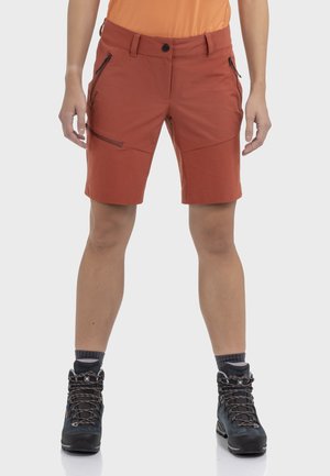 Person trägt rostfarbene Outdoor-Shorts mit mehreren Reißverschlusstaschen und schwarze Wanderschuhe mit grauen Socken.