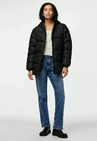 Casaco puffer preto com gola alta, apresentando secções acolchoadas. Usado com um top de tom claro com riscas e jeans azuis, complementado por botas pretas.