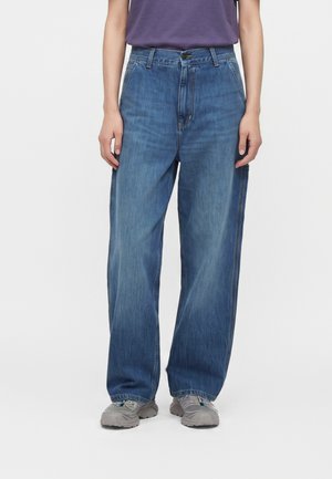 Person iført løstsiddende blå denimjeans, grå sneakers og en lilla skjorte, stående mod en ensfarvet hvid baggrund.